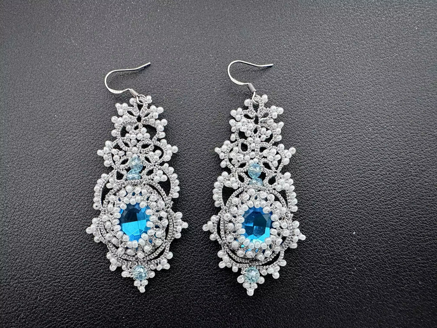 Pair of handcrafted Nebula tatting lace earrings made with shimmering silver thread, white beads, and blue central crystals. Ideal for formal evening wear.
