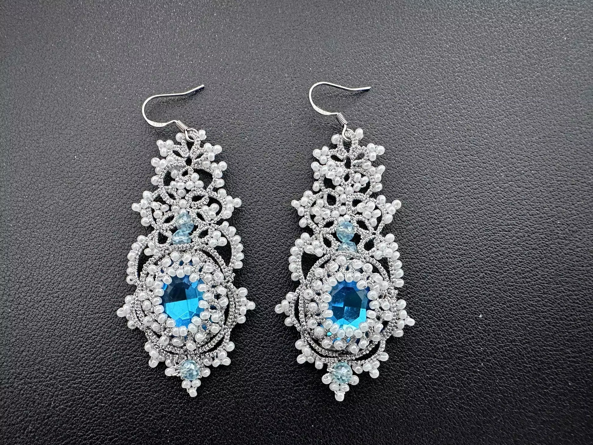 Pair of handcrafted Nebula tatting lace earrings made with shimmering silver thread, white beads, and blue central crystals. Ideal for formal evening wear.